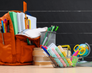 Various school supplies, backpack, pens, pencils, scissors, etc., sitting on desk in front of chalkboard.