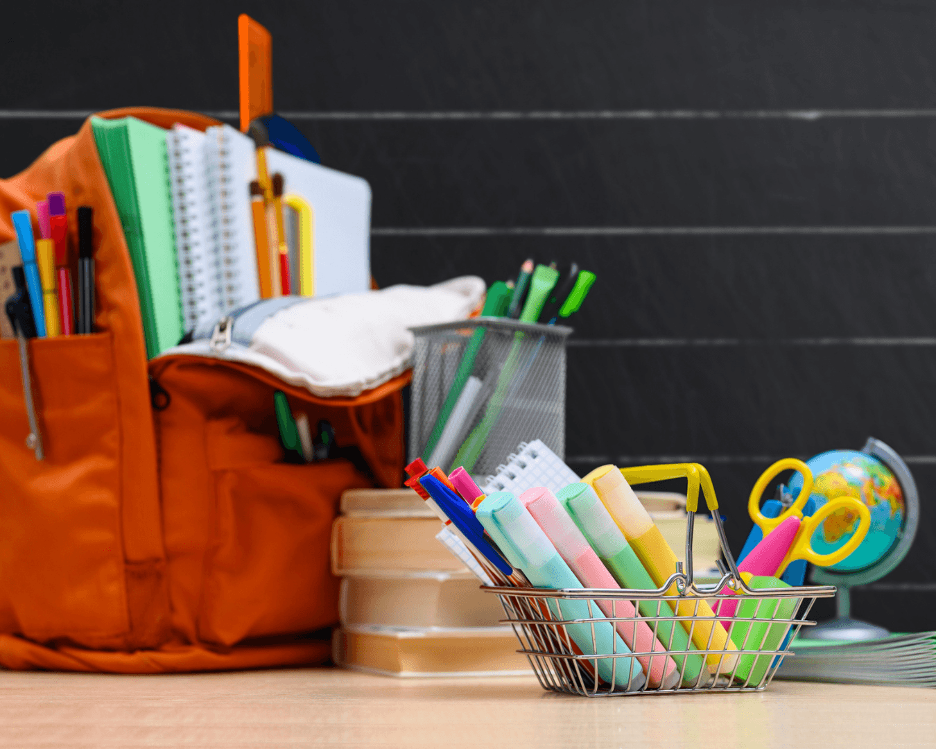 Various school supplies, backpack, pens, pencils, scissors, etc., sitting on desk in front of chalkboard.